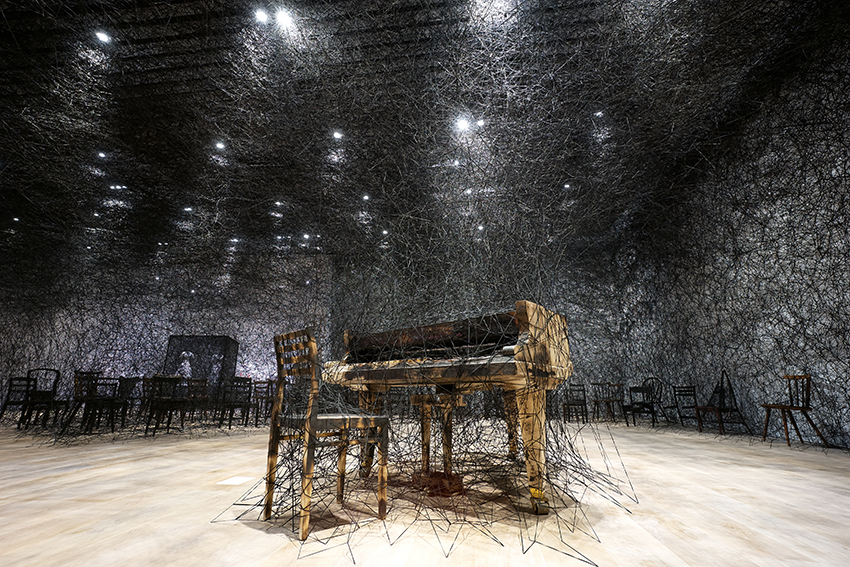塩田千春 | 《靜默中》 燒焦鋼琴、燒焦椅、Alcantara黑線，2002 / 2019年, 尺寸依空間而定  的圖說